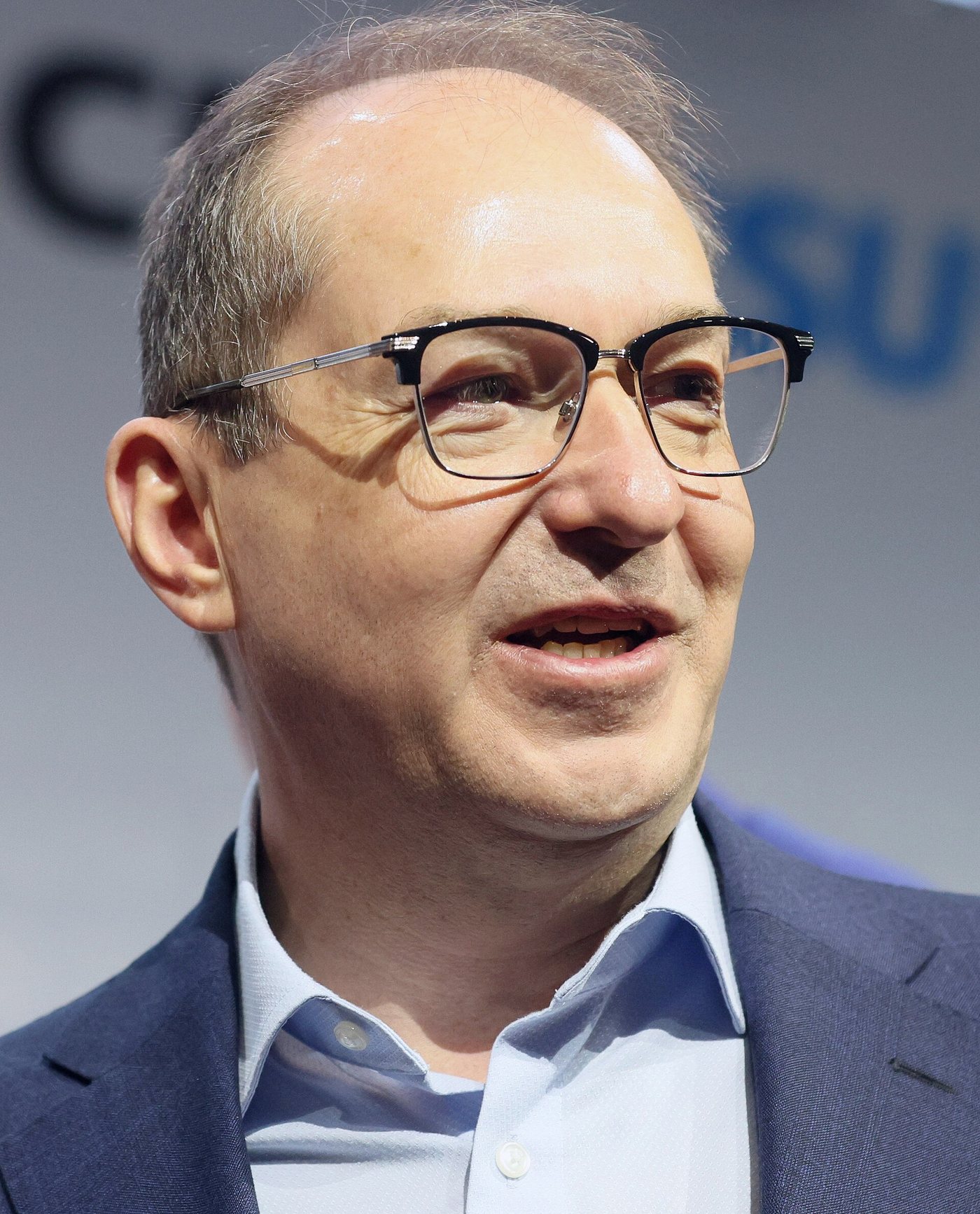 Head-and-shoulders portrait of German Interior Minister Alexander Dobrindt in glasses, a navy jacket and an open-collar light-blue shirt, smiling slightly off-camera against a blurred CSU backdrop.