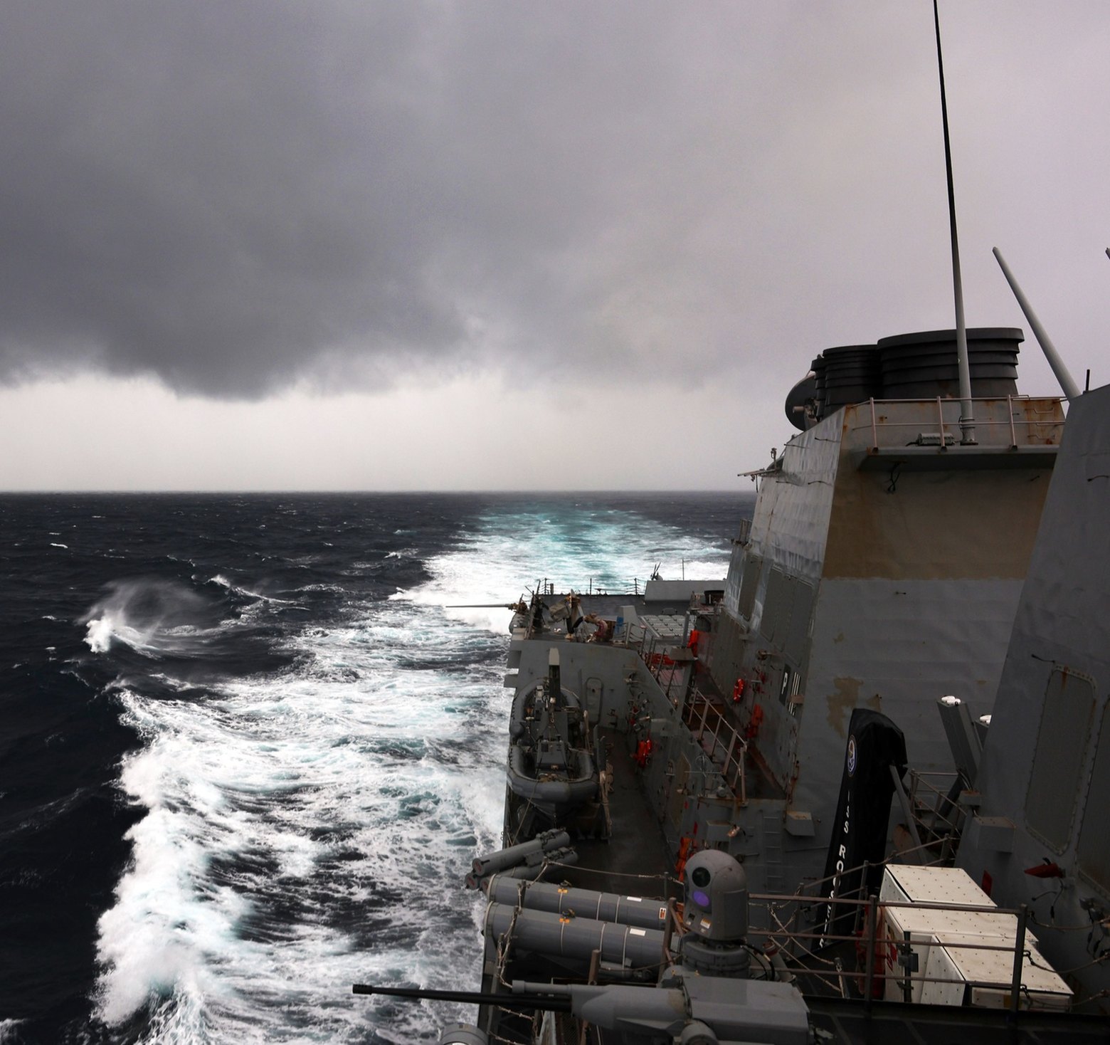 High-angle view from USS Roosevelt (DDG-80) during a Strait of Hormuz transit: grey superstructure and stacks, a stanchion cover reading USS ROOSEVELT, angled missile tubes, a deck gun, orange life rings, and a long white wake on dark choppy water under heavy cloud.